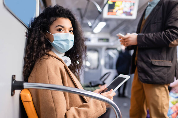 young african american woman in medical mask using digital tablet near man in subway with blurred background 