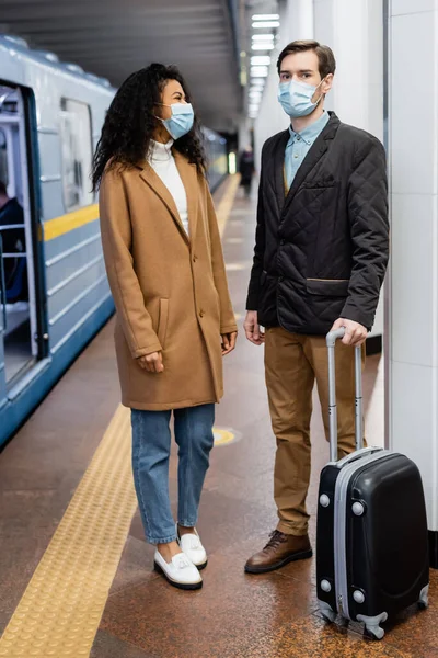 stock image interracial couple in medical masks standing on platform with luggage 