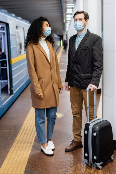 interracial couple in medical masks standing on platform with luggage 