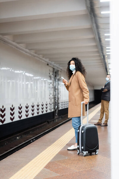 full length of african american woman in medical mask standing with luggage near man in subway 