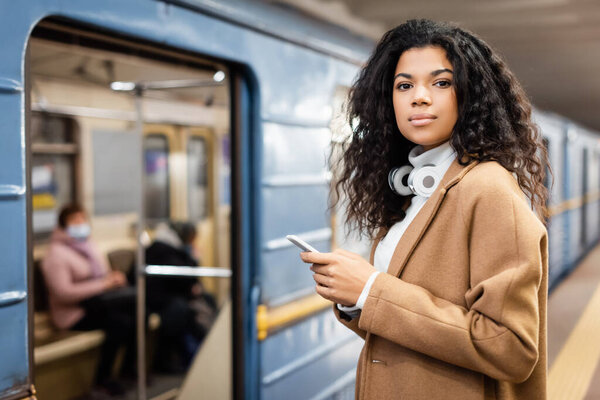 smiling african american woman in wireless headphones holding smartphone in subway 