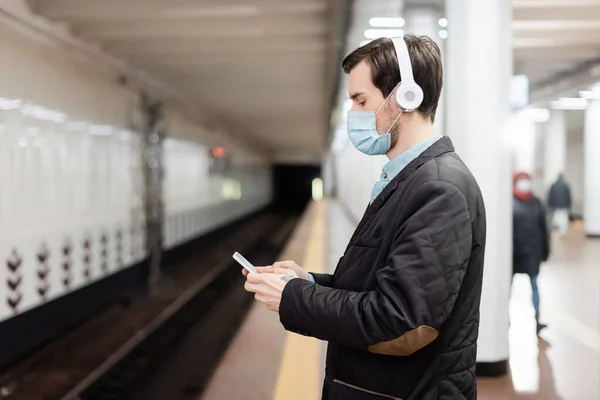 side view of tattooed man in medical mask and wireless headphones holding smartphone in subway