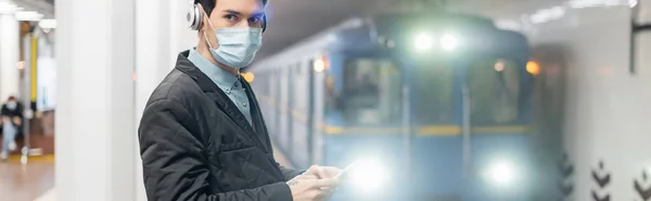 man in medical mask and wireless headphones holding smartphone in subway, banner