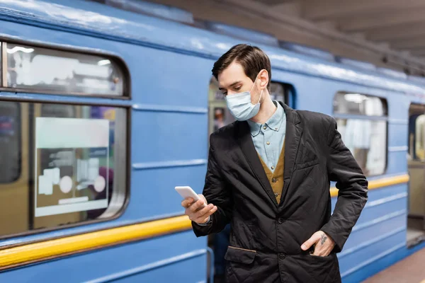 stock image tattooed man in medical mask holding smartphone and standing with hand on pocket near wagon of metro
