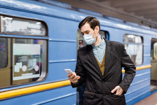 tattooed man in medical mask holding smartphone and standing with hand on pocket near wagon of metro