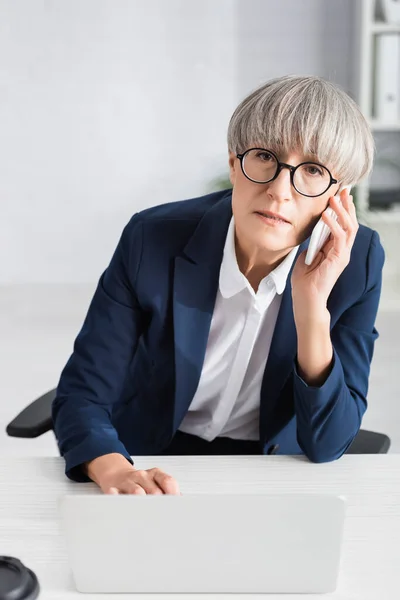Mature Businesswoman Glasses Talking Smartphone While Using Laptop Blurred Foreground — Stock Photo, Image