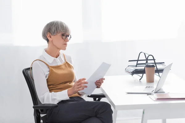 mature team leader sitting at desk and holding documents in office