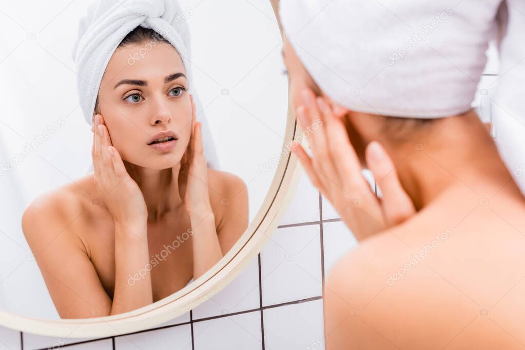 young woman with towel on head touching face while looking in mirror, blurred foreground