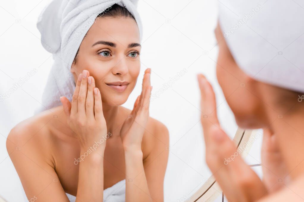 young woman with towel on head applying facial scrub while looking in mirror, blurred foreground