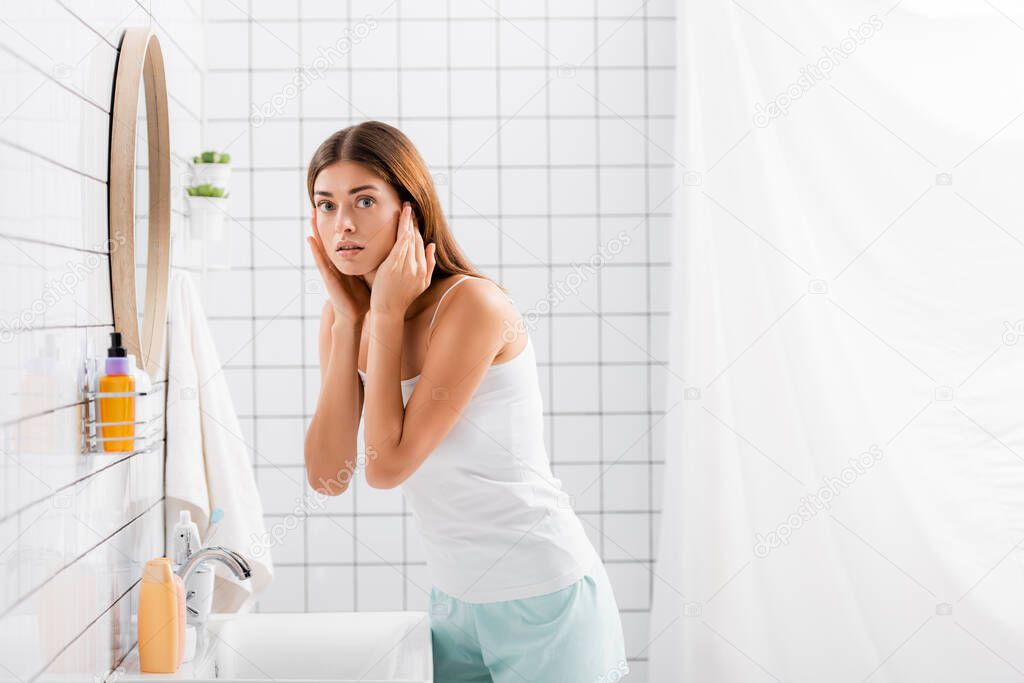 worried young woman in singlet and shorts touching face while looking at camera in bathroom
