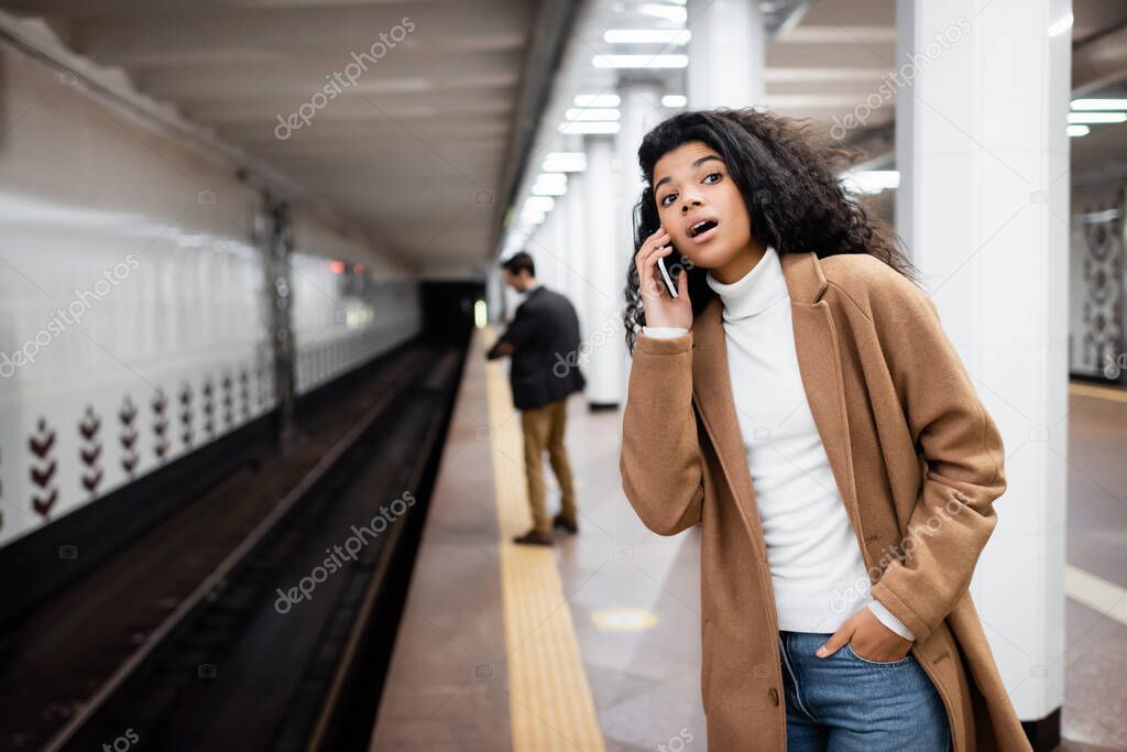 surprised african american woman talking on smartphone and looking away in subway on blurred background