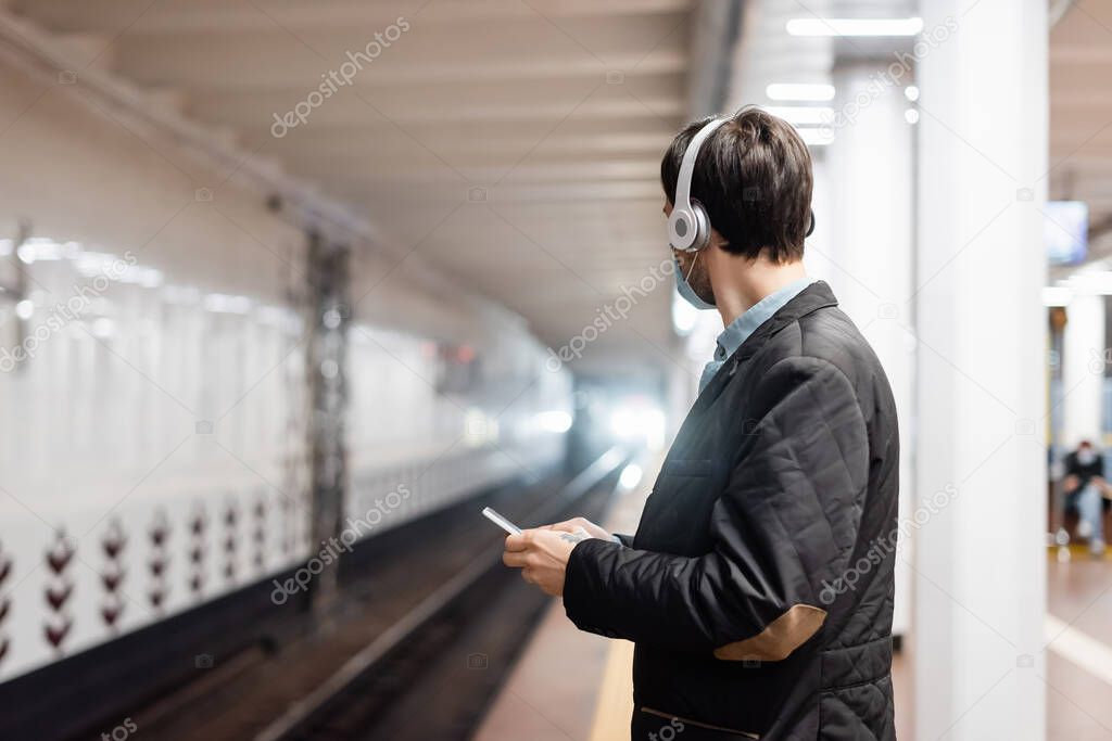 tattooed man in medical mask and wireless headphones holding smartphone in subway 