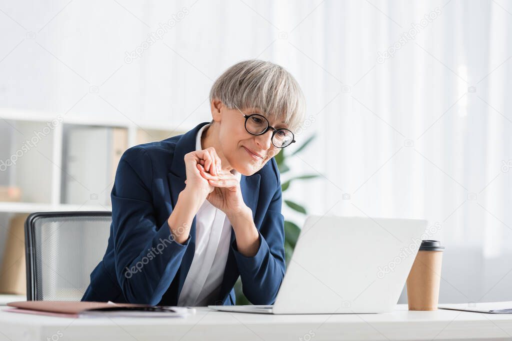 cheerful and mature team leader in glasses looking at laptop 