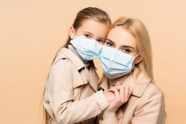 Tıbbi maskeli anne ve kız bejde el ele tutuşuyorlar.