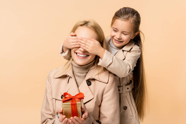 kid covering eyes of happy mother with gift box isolated on beige