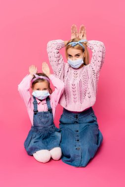 Tıbbi maskeli anne ve kızı pembe ellerle tavşan kulağı yapıyorlar. 