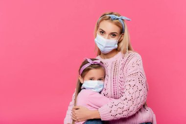 Anne ve kız tıbbi maskeler takıp kameraya bakarken pembe renkte izole edilmiş bir şekilde kucaklaşıyorlar. 