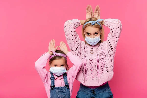 医療用マスクの母親と娘がピンクに手を隔離してウサギの耳を作る — ストック写真