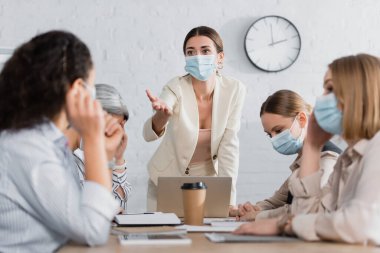 Tıbbi maskeli genç takım lideri bulanık ön planda ırklar arası iş kadınlarını işaret ediyor. 