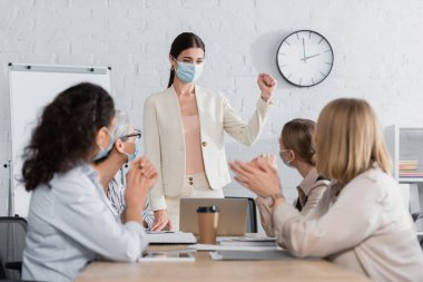 Tıbbi maskeli genç takım lideri yumruklarını sıkmış ırkçı iş kadınlarıyla ön planda bulanık duruyor. 
