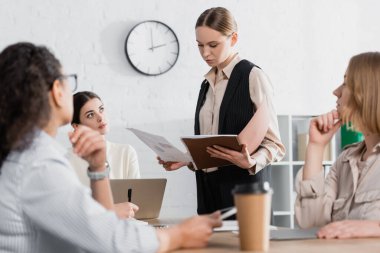 young businesswoman holding folder near team leader and multicultural coworkers during meeting  clipart