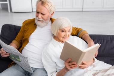 Gülümseyen yaşlı çift evdeki kanepede kitap ve haber okuyor. 