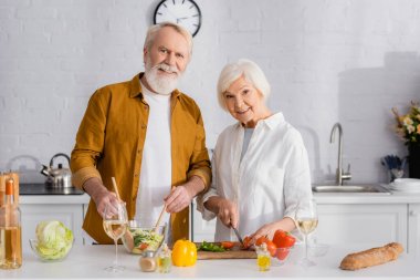 Smiling elderly couple cooking fresh salad near wine and baguette in kitchen  clipart
