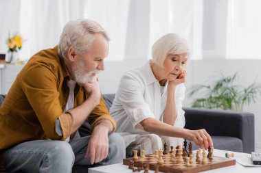 Yaşlı kadın evde düşünceli kocasının yanında satranç oynuyor. 