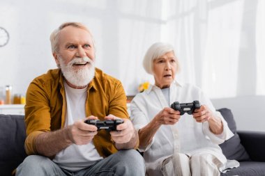 KYIV, UKRAINE - DECEMBER 17, 2020: Cheerful senior man playing video game near upset wife on blurred background  clipart
