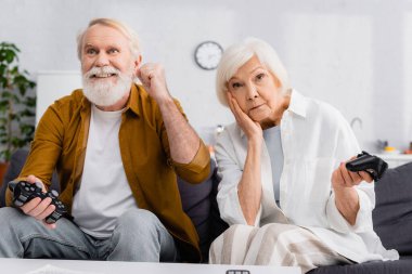 KYIV, UKRAINE - 17 Aralık 2020: heyecanlı yaşlı adam evde üzgün karısının yanında joystick tutuyor 