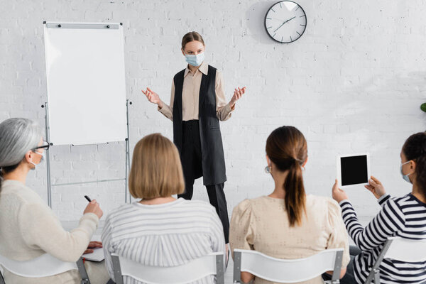 оратор в медицинской маске стоя и жесты, глядя на межрасовых деловых женщин на размытом переднем плане