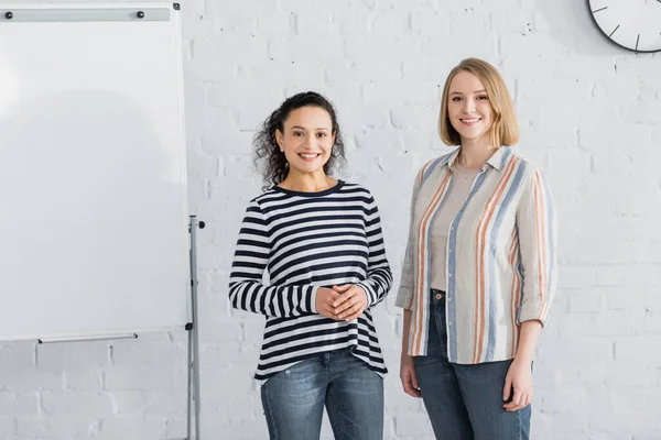 african american businesswoman smiling near colleague and flipchart in meeting room