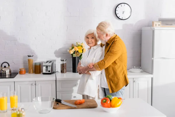 Homem Idoso Abraçando Esposa Sorridente Perto Tábua Corte Legumes Frescos — Fotografia de Stock