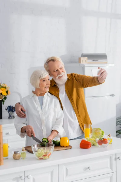上了年纪的夫妇在厨房里的橙汁边做饭 一边自拍 — 图库照片