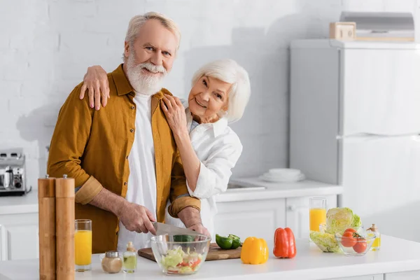 Mujer Sonriente Abrazando Marido Mayor Cortando Verduras Cerca Jugo Naranja — Foto de Stock