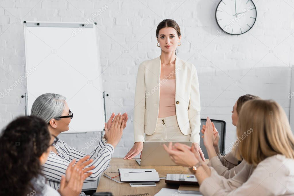 confident team leader looking at camera while multicultural audience applauding on blurred foreground 