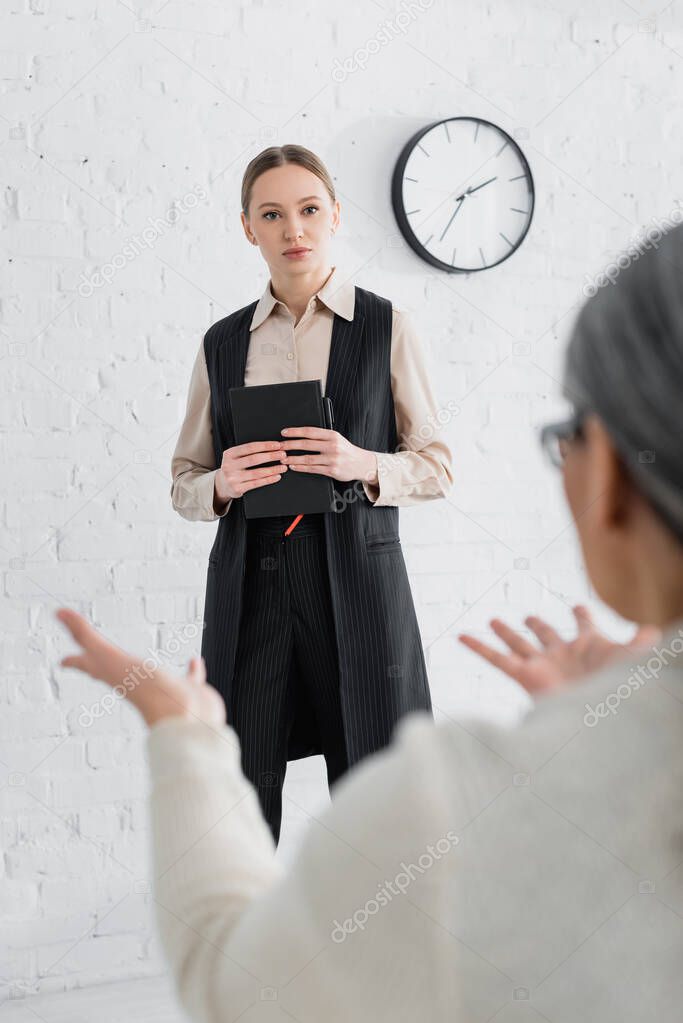 speaker standing with notebook and looking at businesswoman gesturing on blurred foreground during lecture