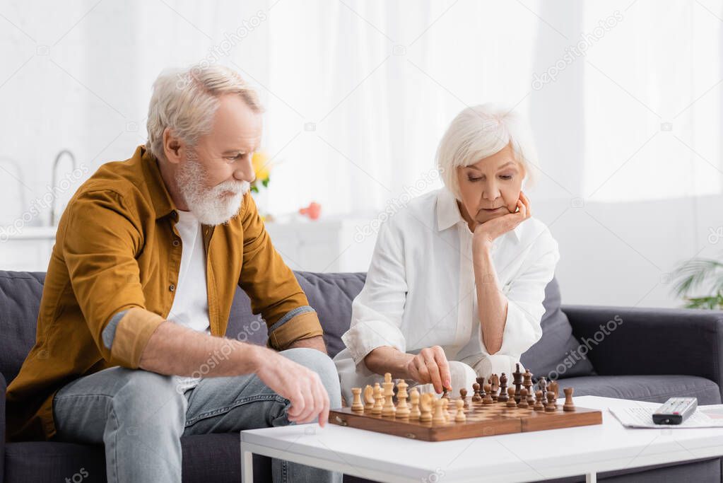 Pensive elderly couple playing chess near remote controller on blurred background 