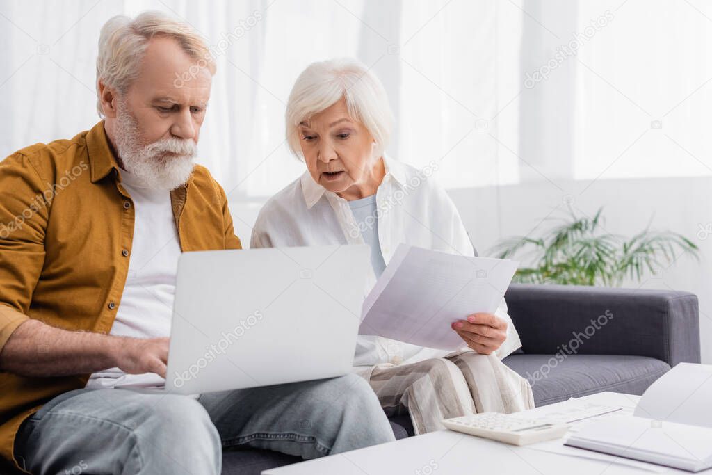 Senior man using laptop near wife with papers, calculator and notebook on table 