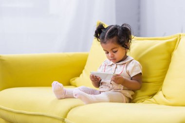 Küçük Afrikalı Amerikalı kız kanepede otururken akıllı telefon kullanıyor.