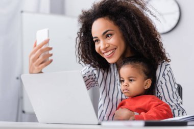 Mutlu Afro-Amerikan annesi kızıyla birlikte bulanık ön planda dizüstü bilgisayarın yanında selfie çekiyor.