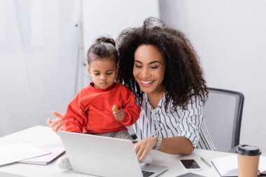 Küçük Afrikalı Amerikalı çocuk elinde lezzetli kurabiyelerle gülümseyen annesinin yanında oturuyor ve dizüstü bilgisayarına bakıyor.