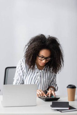 Gözlüklü Afrikalı Amerikalı hesap makinesi kullanarak masadaki laptopu ve kağıt bardağı sayıyor.