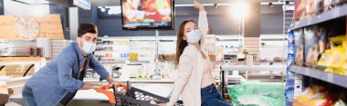 Man in medical mask rolling shopping trolley with girlfriend in supermarket on blurred foreground, banner 