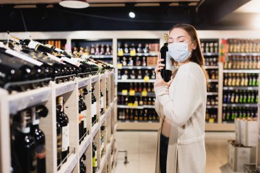 Tıp maskeli bir kadın süpermarkette şarap şişesi tutuyor. 