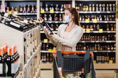 Tıbbi maskeli bir kadın markette alışveriş arabasının yanında şarap şişesi tutuyor. 
