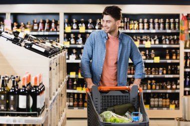 Süpermarkette alışveriş arabasının yanında şarap şişelerine bakan gülümseyen adam. 