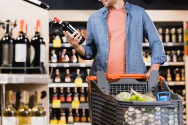 Cropped view of smiling man holding bottle of wine near shopping cart on blurred foreground in supermarket  clipart