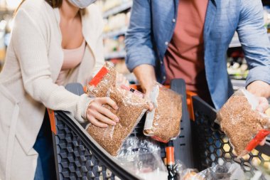Cropped view of couple putting packages with buckwheat in shopping cart on blurred foreground in store  clipart