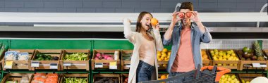 Young couple holding fruits near shopping cart in supermarket, banner 